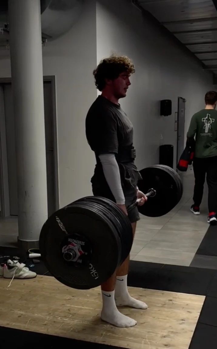 A lifter in socks standing tall at the top of a deadlift, the bar loaded with black plates