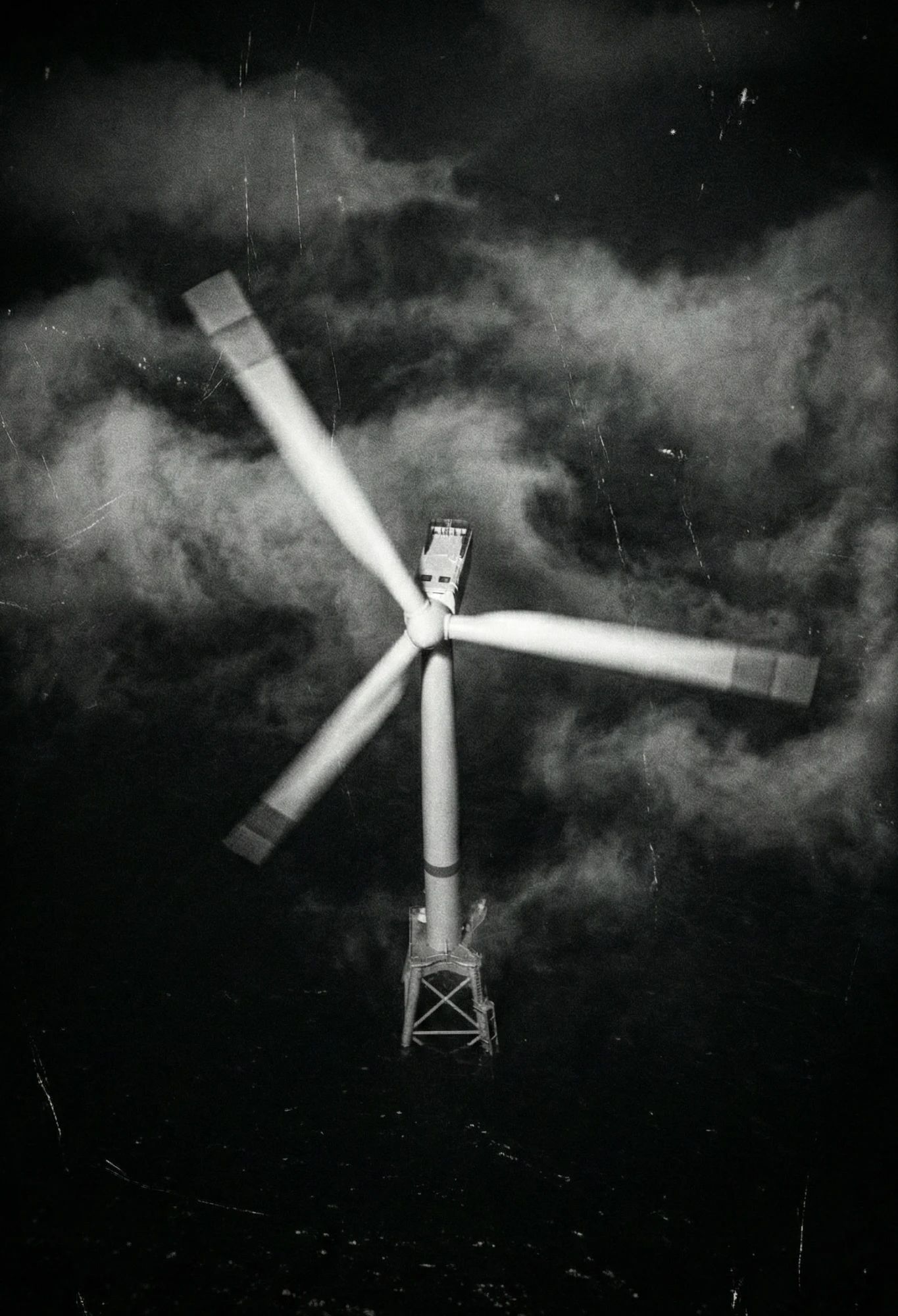 A wind turbine at sea from above, its blades blurred, on scratched black and white film